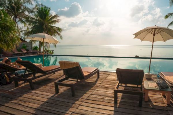A serene poolside view featuring lounge chairs, umbrellas, palm trees, and a calm sea under a partially cloudy sky.