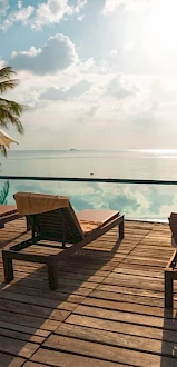 A wooden deck with sun loungers by an infinity pool, palm trees, and ocean view, under a bright sky.