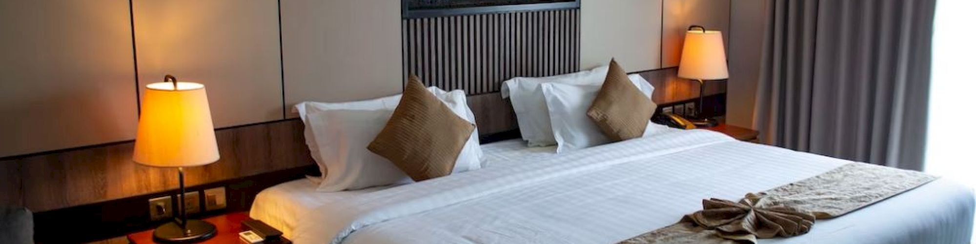 A neatly arranged hotel room features a large bed, decorative pillows, warm lighting, and curtain-covered windows near a seating area.