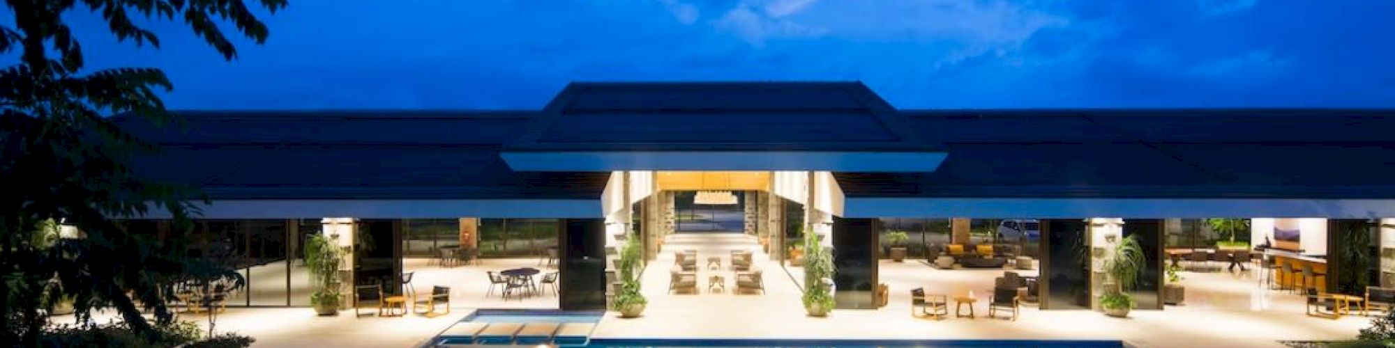 The image shows an upscale building with an illuminated pool at night, surrounded by lounge chairs and trees under a deep blue sky.