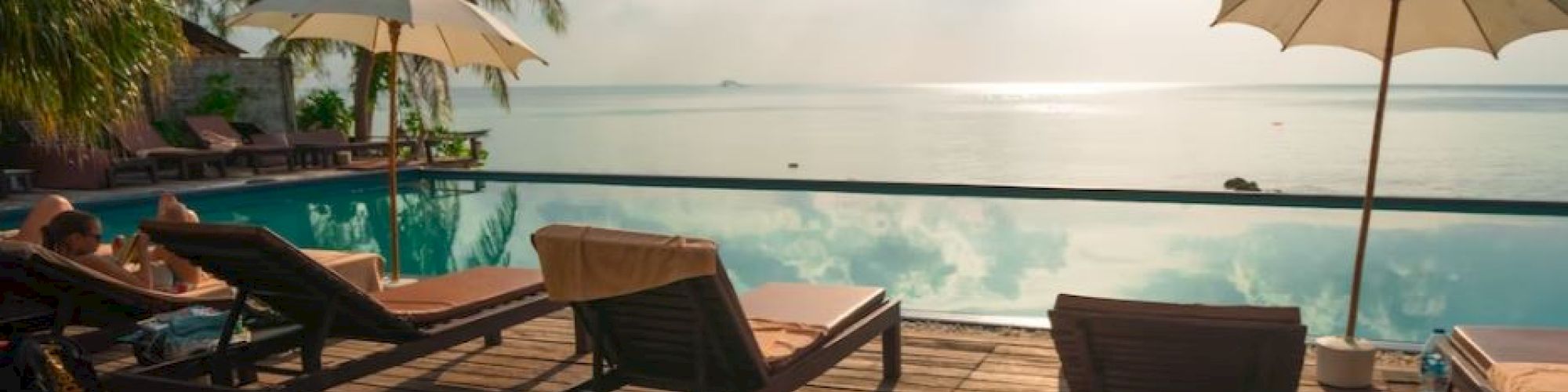 A serene poolside view with lounge chairs, umbrellas, and palm trees overlooking a calm water body under a partly cloudy sky.