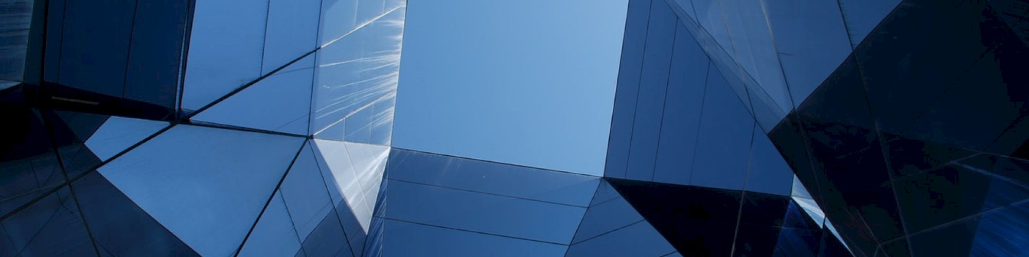 A view of a geometric glass and steel structure, looking upward to a clear blue sky framed by reflective building surfaces.