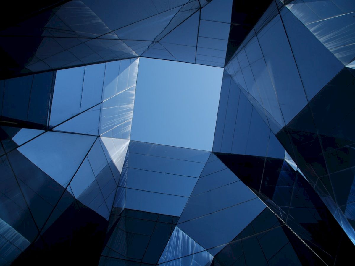 The image shows a geometric architectural structure with reflective glass surfaces forming a skylight, revealing the blue sky above.