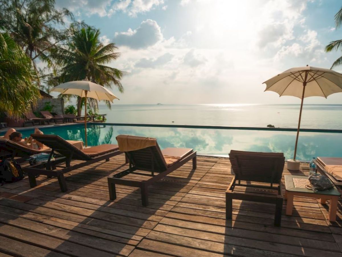 A serene poolside deck with loungers and umbrellas overlooks a calm sea under a partly cloudy sky, with lush greenery nearby.