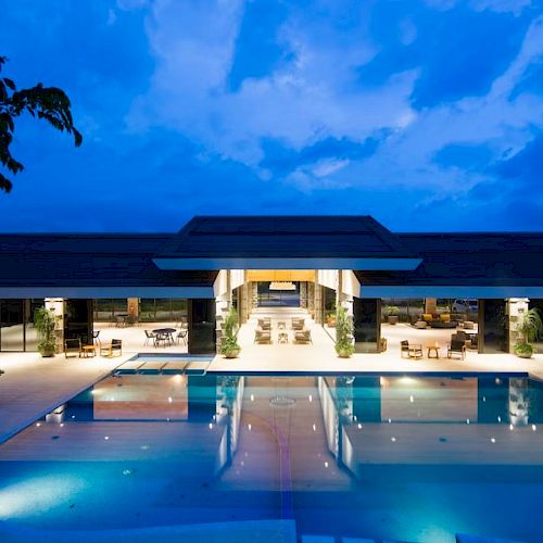 A modern building with large glass windows and a spacious illuminated pool against a blue evening sky completes the view.
