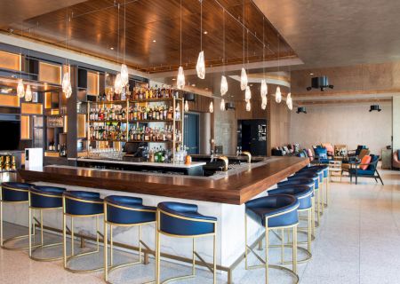 A modern, stylish bar with blue chairs, pendant lights, and a fully stocked shelves, surrounded by a cozy seating area.
