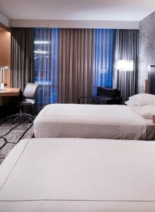 The image shows a modern hotel room with two beds, a wall-mounted TV, a desk with a chair, and contemporary decor.