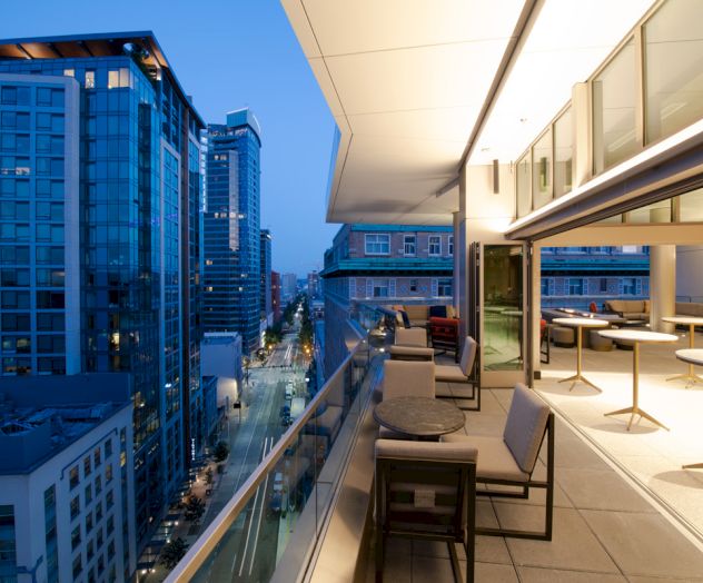 A modern balcony overlooking a cityscape with tall buildings at dusk, featuring outdoor seating and a bright, open indoor space.