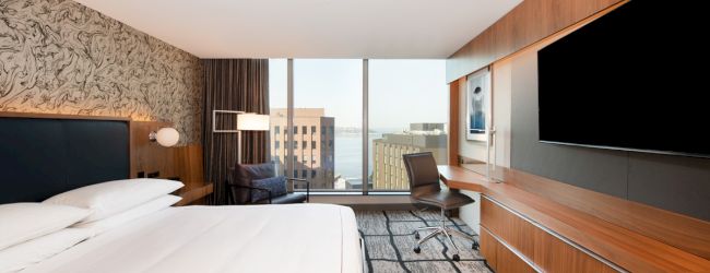 A modern hotel room with a bed, wall-mounted TV, desk, chair, and large window offering a city view ending the scene.