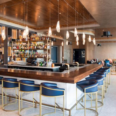 A modern bar with a central counter, shelves of bottles, blue stools, pendant lights, and a lounge area in the background.