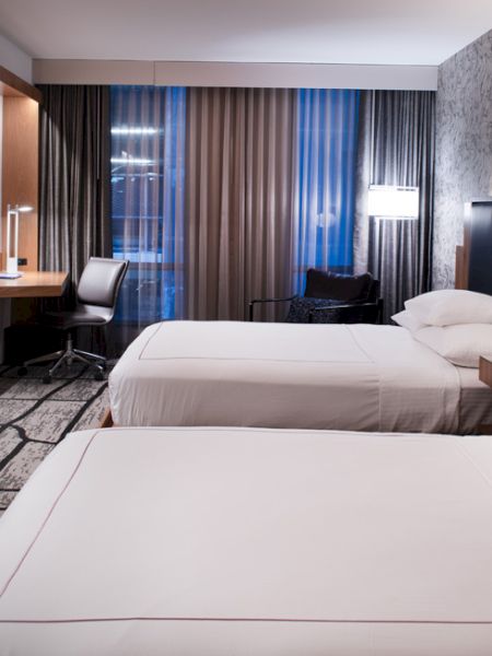 A modern hotel room with two beds, a flat-screen TV, desk, chair, and stylish decor, featuring patterned wallpaper and carpet.