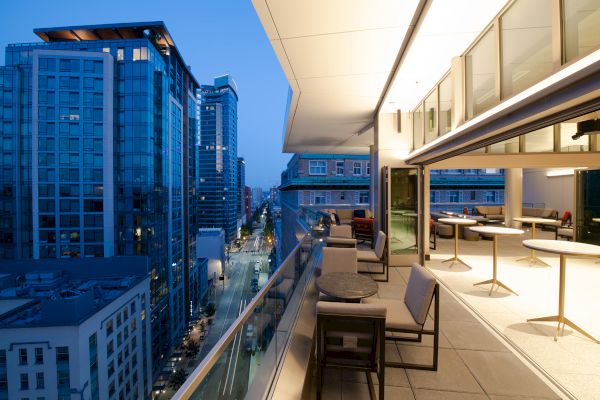 A modern rooftop terrace overlooks a city street, offering tables and seating with a view of adjacent high-rise buildings at dusk.