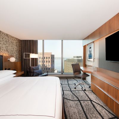 A modern hotel room with a large bed, desk, chair, and a big TV. There’s a window with a city and water view.