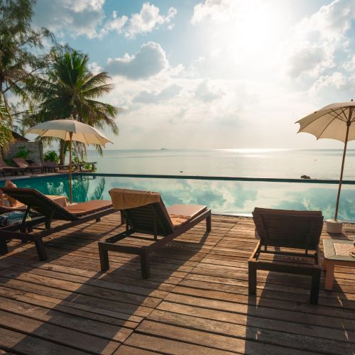 Sun loungers and umbrellas by an infinity pool, overlooking a serene ocean view with palm trees under a bright sky.