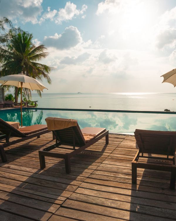 Sun loungers and umbrellas by an infinity pool, overlooking a serene ocean view with palm trees under a bright sky.