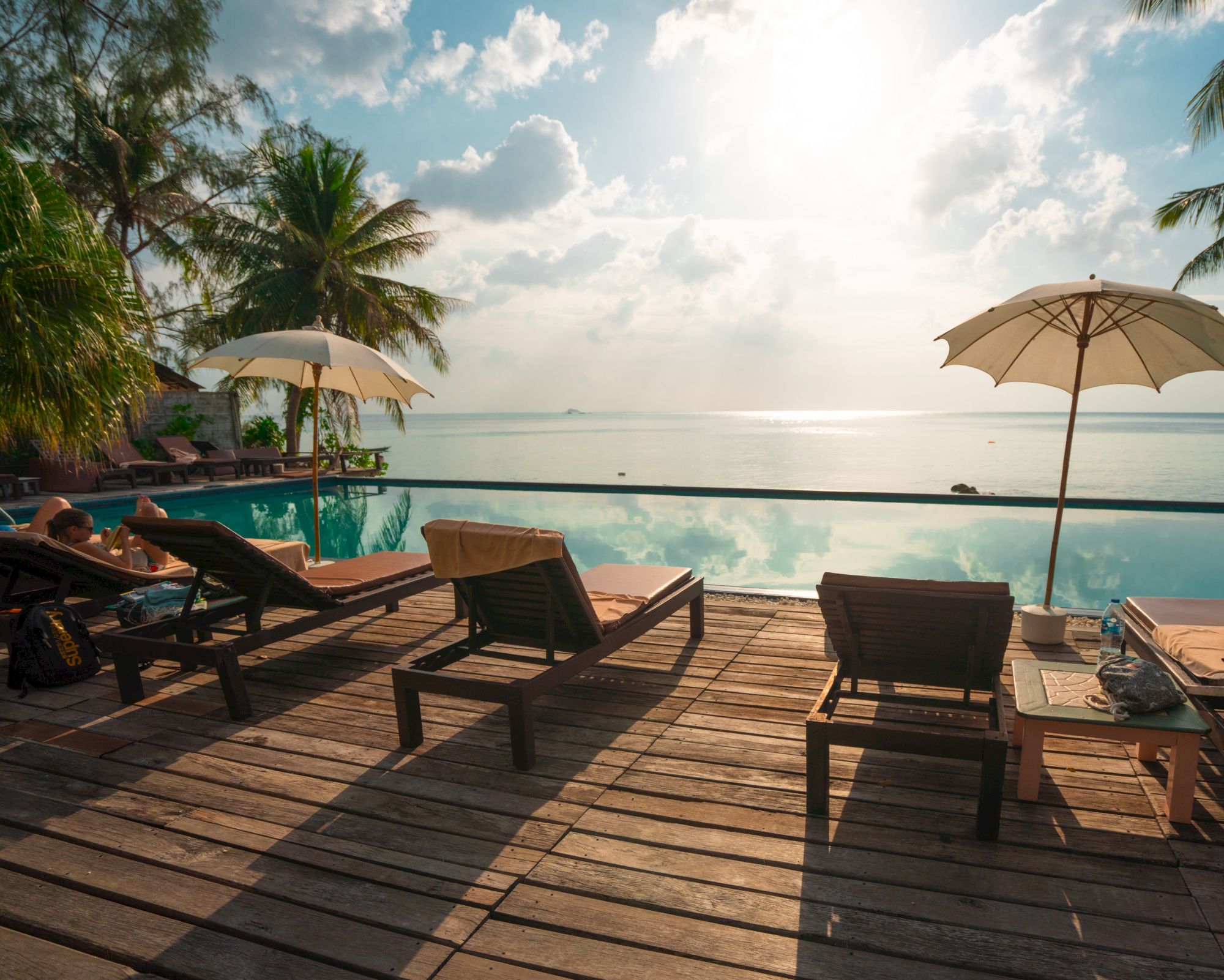 A serene beach scene with lounge chairs, umbrellas, and a pool overlooking the ocean, framed by palm trees under a partly cloudy sky.