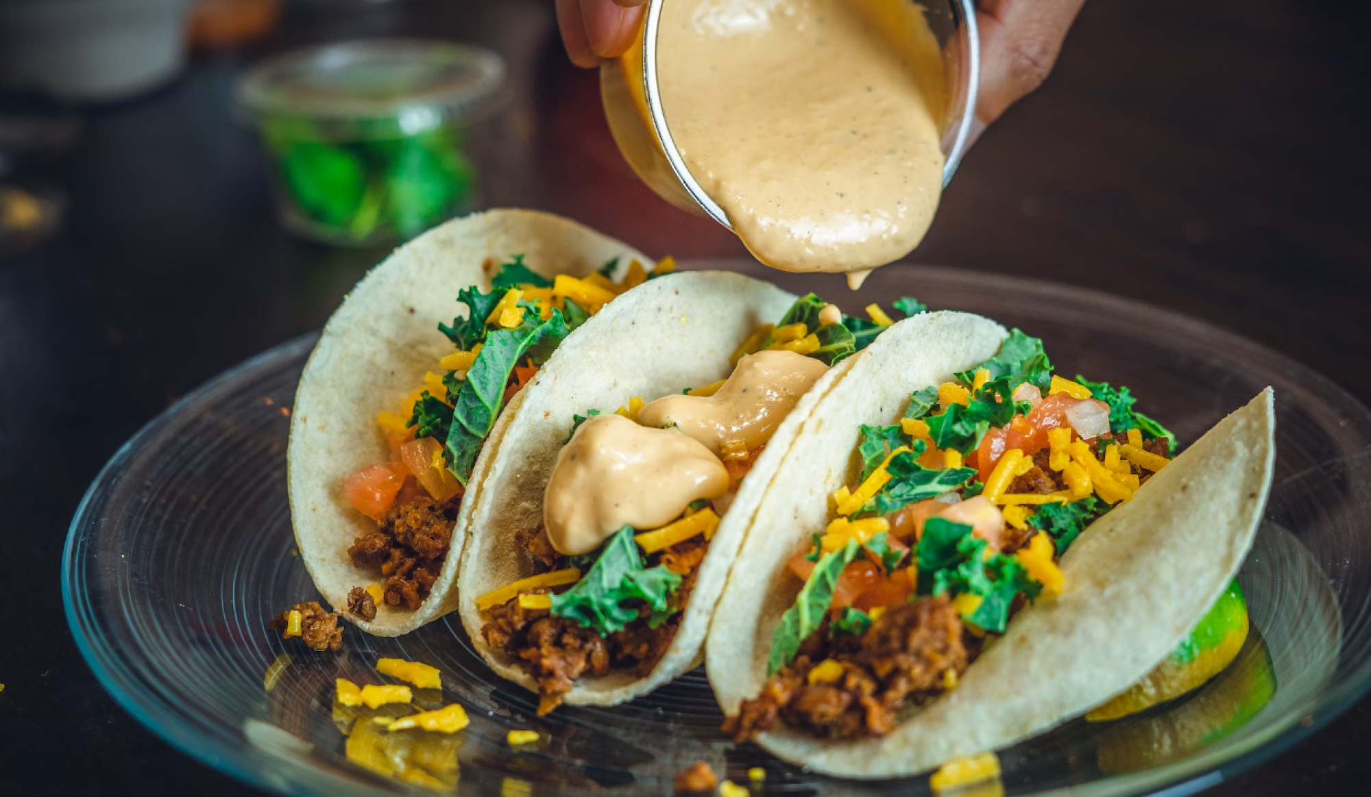 Three tacos with meat, cheese, kale, and tomatoes are being topped with a creamy sauce on a glass plate.