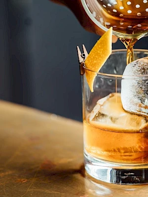 A hand pours a brown liquid into a glass with ice and a citrus peel garnish on a wooden surface.