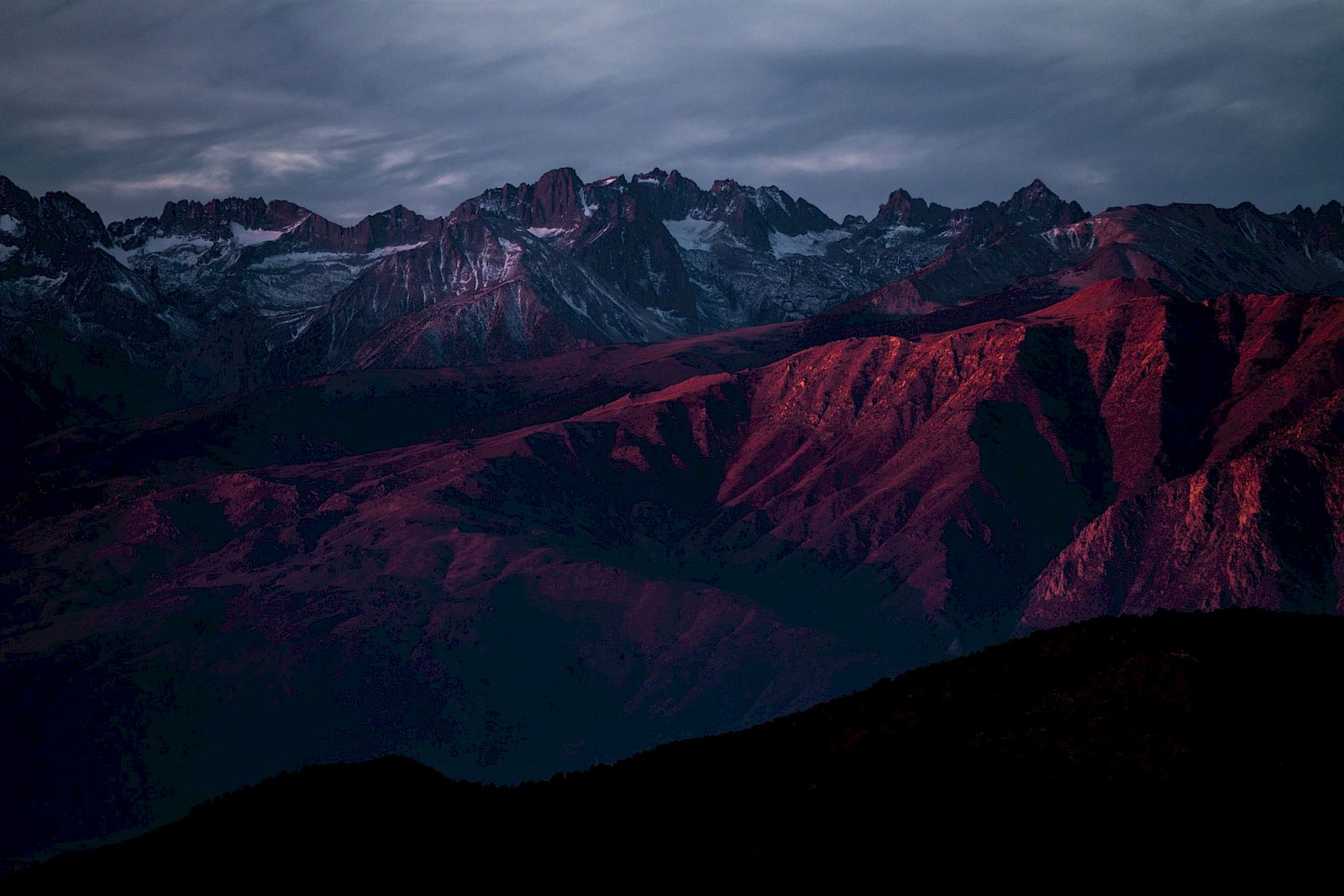 A mountain range bathed in deep red light under a cloudy sky.