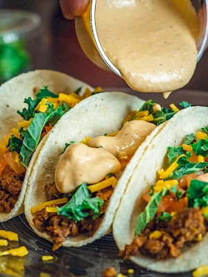 Three tacos filled with ground meat, cheese, lettuce, and tomatoes are on a plate, with sauce being poured over them.