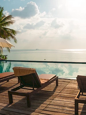 Lounge chairs by a pool overlook a serene ocean, with palm trees and umbrellas providing shade under a partly cloudy sky.