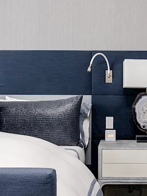 The image shows a bedroom with a blue headboard, white bedding, blue pillow, and a bedside table with a lamp and wall-mounted reading light.