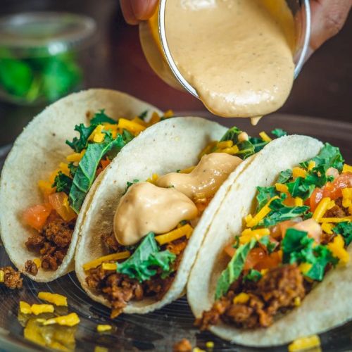 Three tacos with ground beef, lettuce, tomatoes, and cheese are on a plate. A hand is pouring sauce over them.