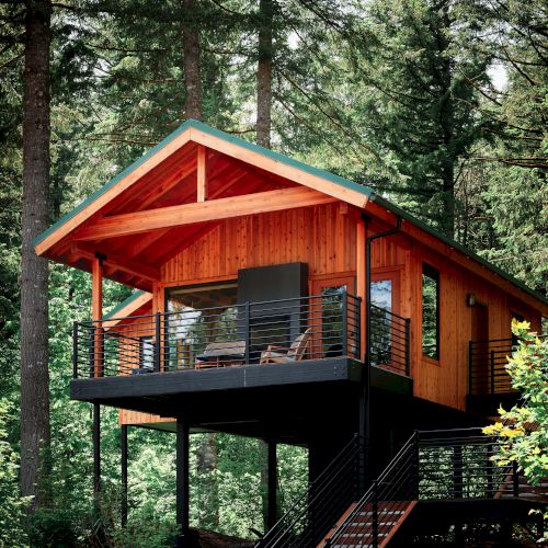 A modern wooden cabin with a balcony sits elevated among tall trees, surrounded by green foliage in a forest setting.