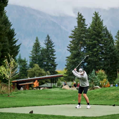 A golfer swings on a lush course surrounded by trees and mountains in the background.