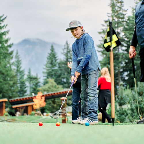 A person playing golf on a green with colorful golf balls and other people around, set against a scenic backdrop of trees and mountains.