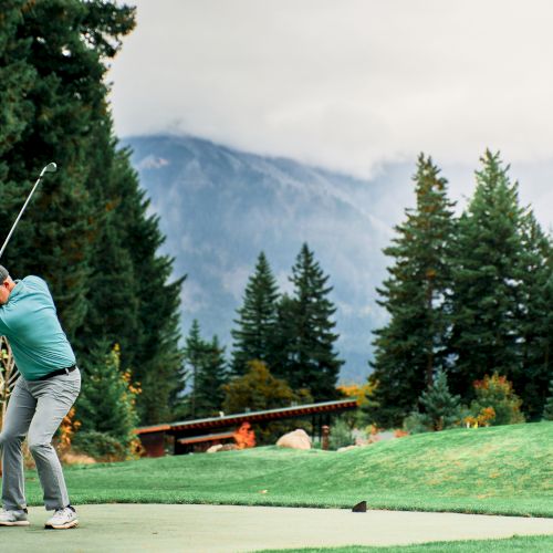 A golfer swings at a ball on a lush course, surrounded by trees with mountains in the background.