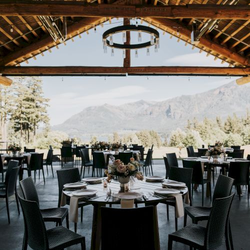 An elegantly set outdoor dining area with round tables and chairs overlooks a scenic mountain landscape under a rustic wooden ceiling.