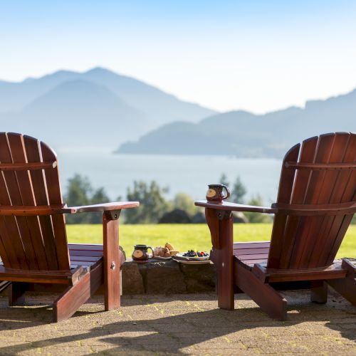 Two wooden chairs overlook a scenic view of mountains and a lake, with mugs and snacks placed between them on a stone ledge.