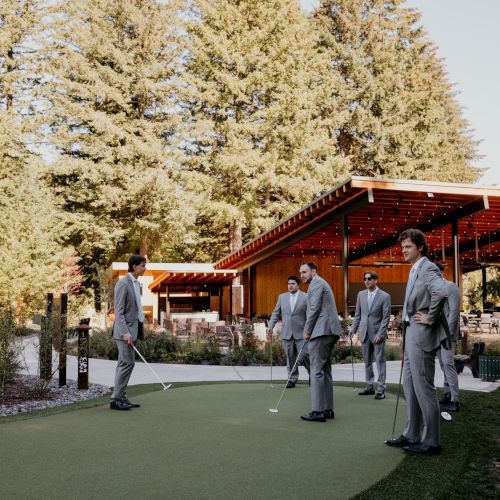 A group of people in suits are standing with golf clubs on a grassy area near a modern wooden building, surrounded by tall trees.