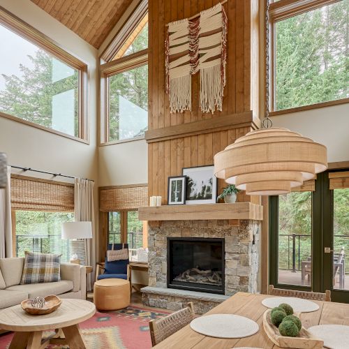 Cozy living room with wood paneling, stone fireplace, large windows, dining table, and cozy seating. Scandinavian decor accents throughout.