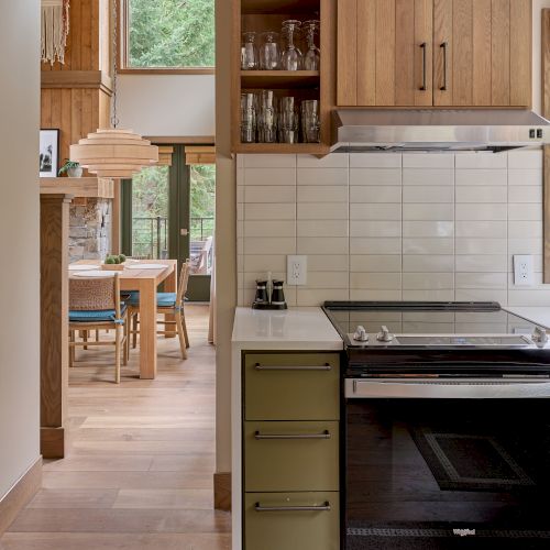 A kitchen with wooden cabinets, open shelving, glassware, an oven, and a view into a cozy dining area with a wooden table and chairs.