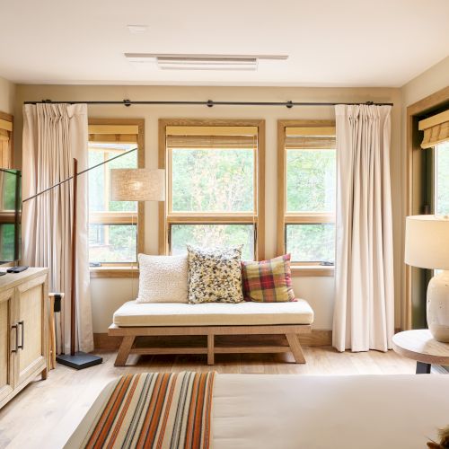 A cozy bedroom with a bed, wooden furniture, TV, floor lamp, and pillows on a bench, framed by large windows with curtains.