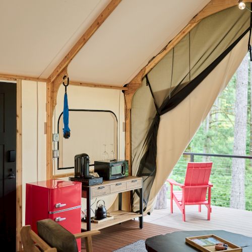 Cozy indoor space with wooden furniture, red fridge, coffee maker, microwave, and string lights. Open to a deck with a red chair.