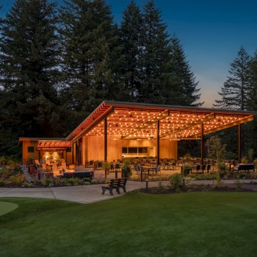 A well-lit open pavilion with string lights, surrounded by trees and greenery, featuring outdoor seating and a partial view of a golf course.