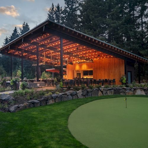 A lit outdoor pavilion with seating set in a lush, wooded area next to a landscaped lawn and putting green.