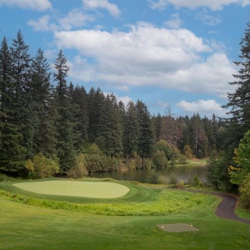 A golf course surrounded by tall trees and a small body of water under a blue sky with clouds.