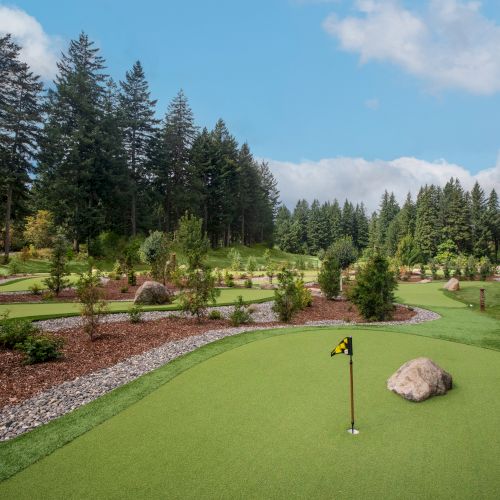 A scenic mini-golf course surrounded by lush greenery and trees, with a hole, flag, and boulder in the foreground under a blue sky.