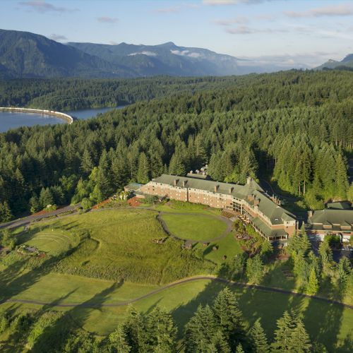 Aerial view of a large building surrounded by lush green forests and mountains, with a river or lake visible in the distance.