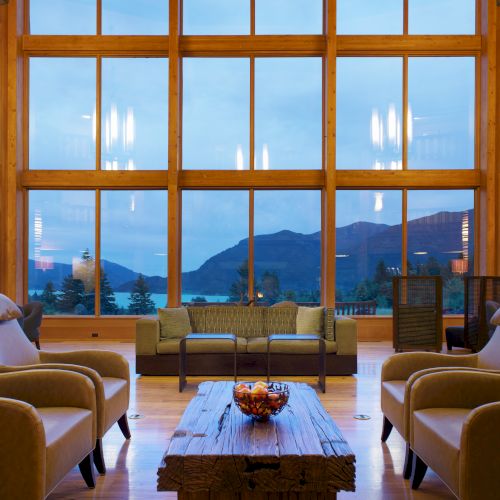 The image shows a cozy, modern living room with large windows, a wooden coffee table, armchairs, and a view of mountains and trees.