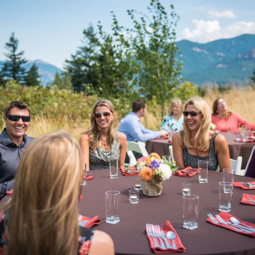 A group of people sits around tables outdoors, enjoying a gathering with a scenic mountain view, under sunny skies.