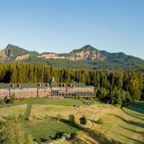 A large building is surrounded by green forests and mountains under a clear blue sky.
