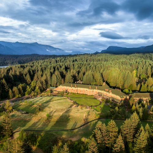 The image shows a large building surrounded by dense forest, with mountains and a body of water in the background under a cloudy sky.