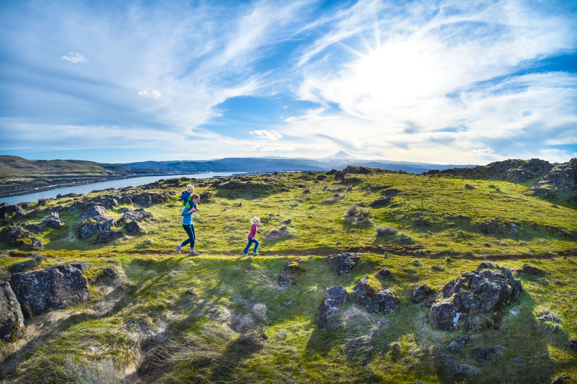 A person and two children walk on a grassy hill with rocks, under a bright sky by a river or lake.