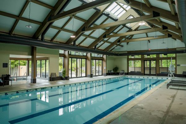 An indoor swimming pool with a high ceiling, large windows, and seating areas, surrounded by wooden beams and natural lighting.