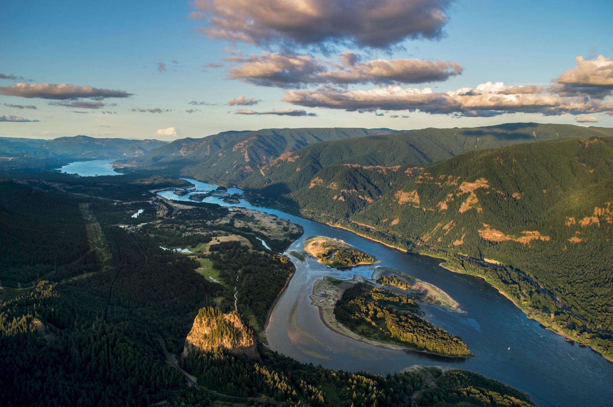 A winding river flows through a lush, mountainous landscape under a partly cloudy sky, illuminated by warm sunlight.